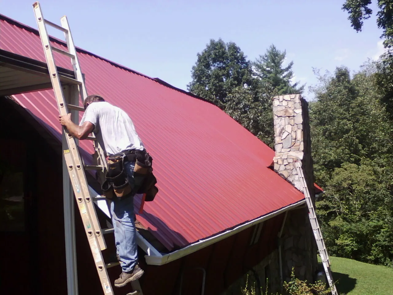 Roofer on a ladder beside a red metal roof and stone chimney — Soffit & Fascia Repair by Roofing Brothers in Pearl
